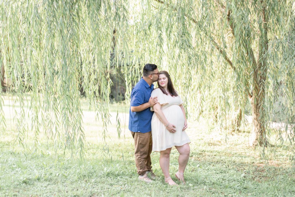 man kisses pregnant wife on the forehead under a willow tree at airfield falls in fort worth