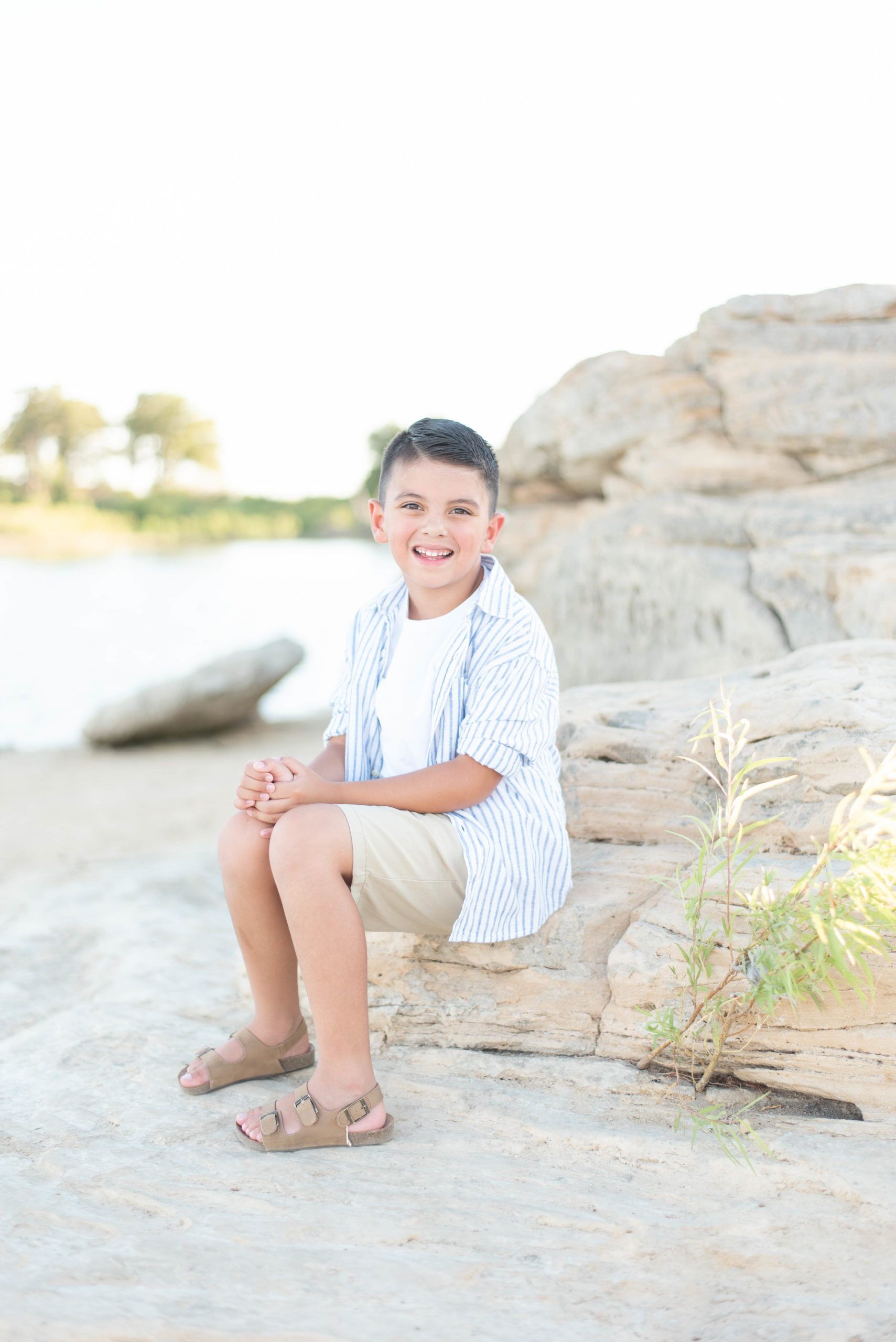 young boy smiles while sitting on a rock at the lake