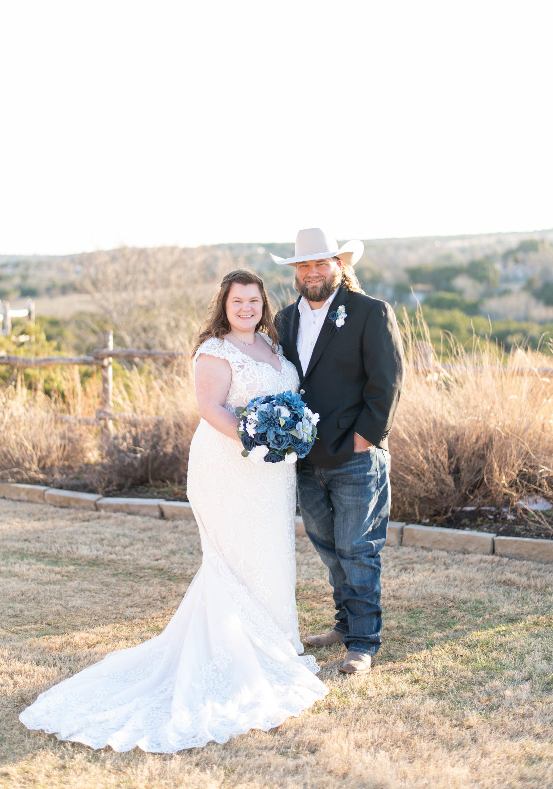 bride and groom are both smiling at the camera holding a blue bouquet with white and blue florals as the sun sets behind them