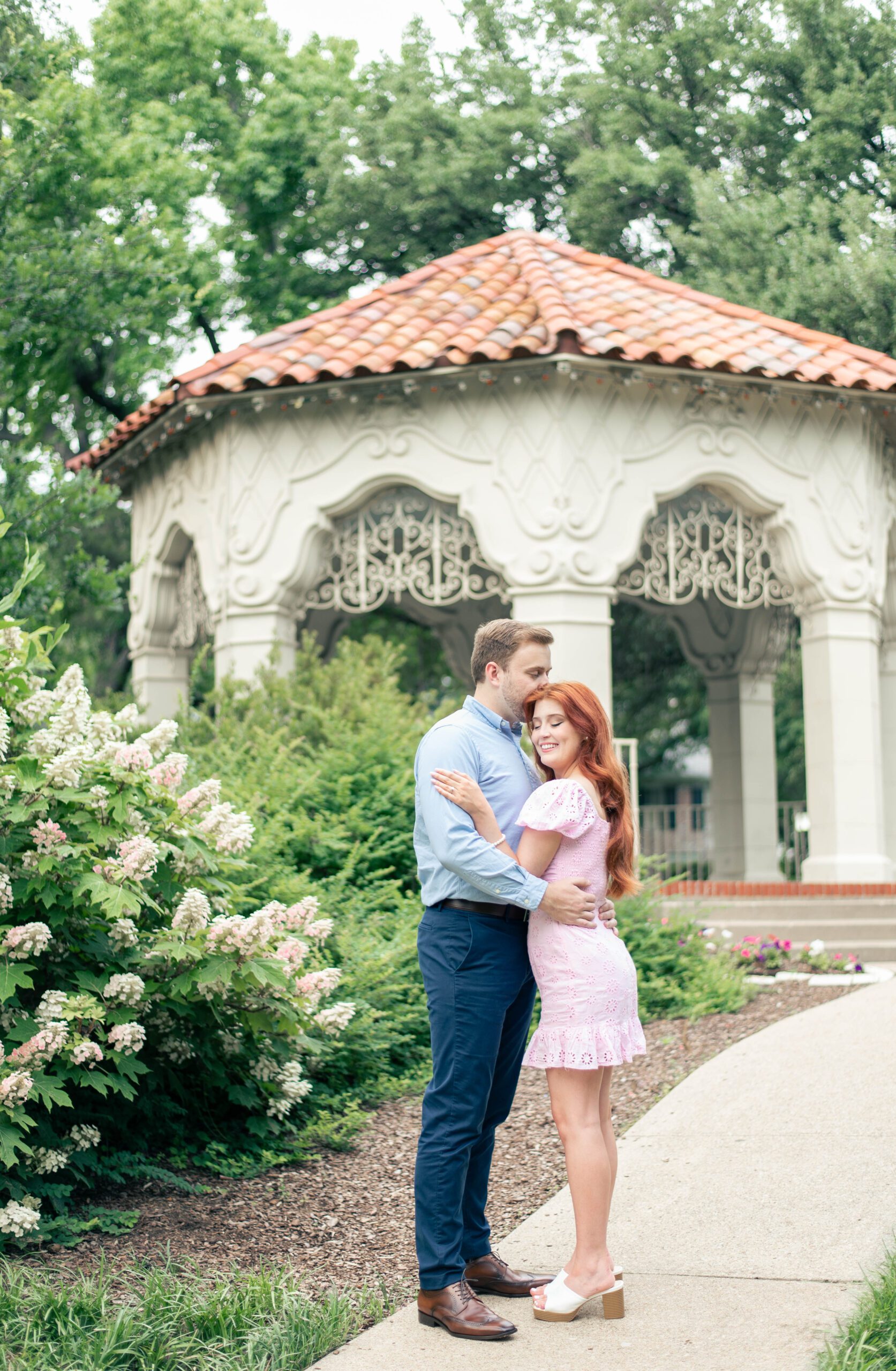 man kisses woman on top of her head while she smiles brightly. Surrounded by lovely flowers and an elegant gazebo at Flippen Park located in Dallas, TX.