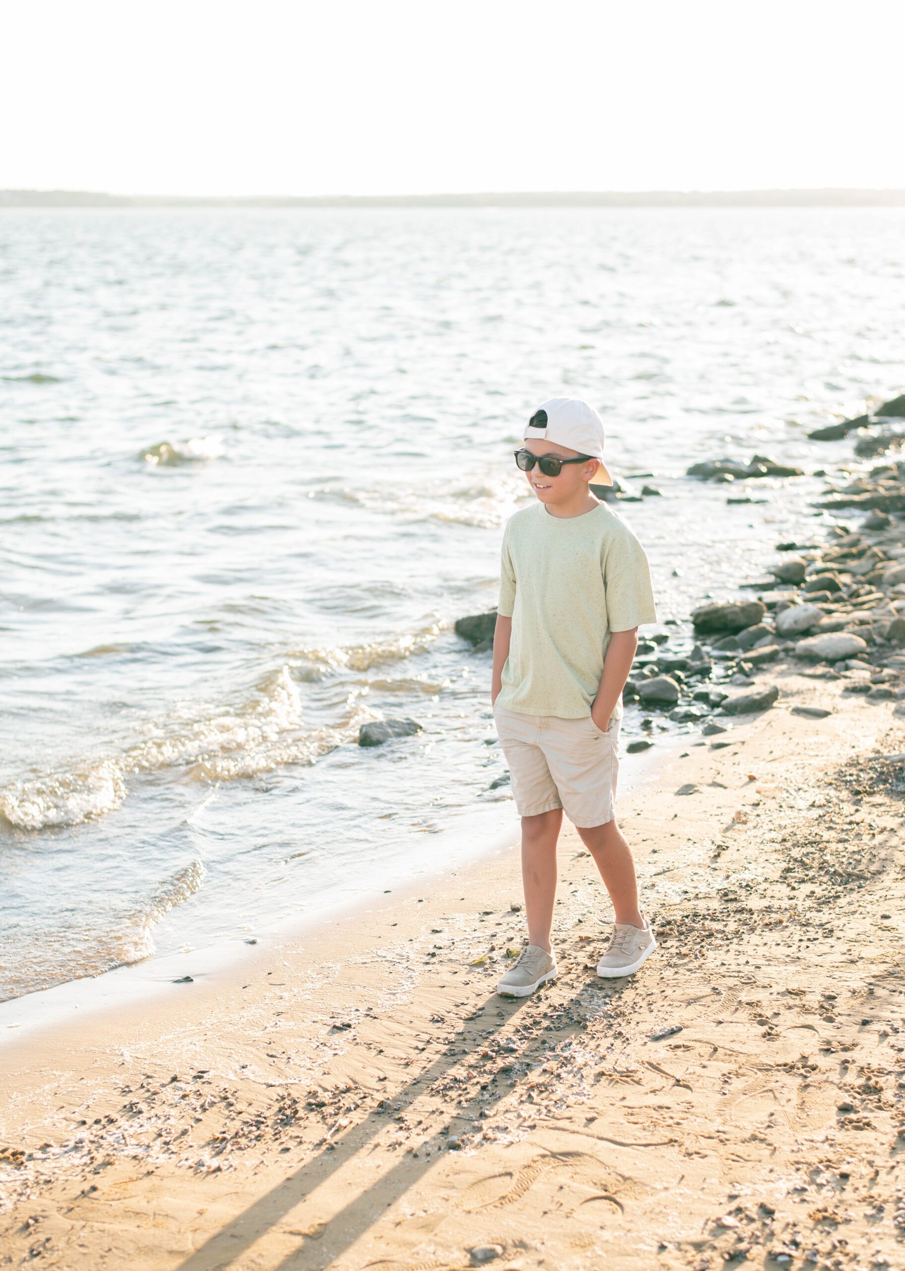 boy walks on the beach with sunglasses on and a backwards baseball cap