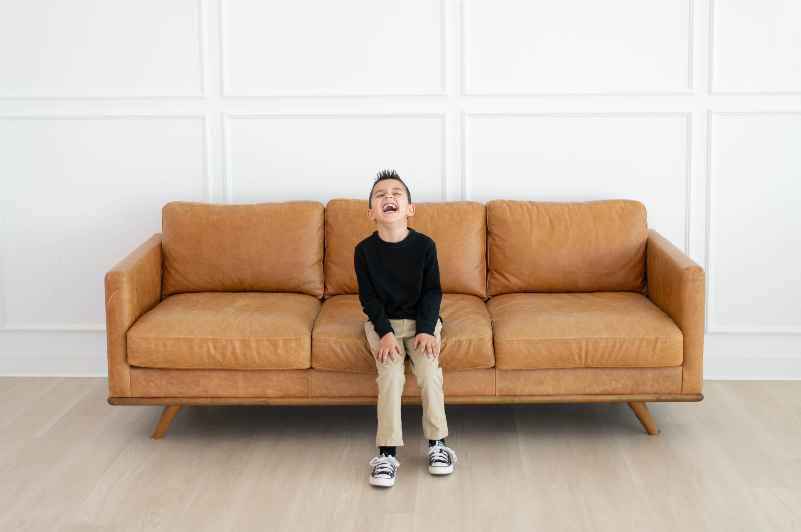 five year old boy sits on a brown leather sofa laughing with his eyes closed and head thrown back