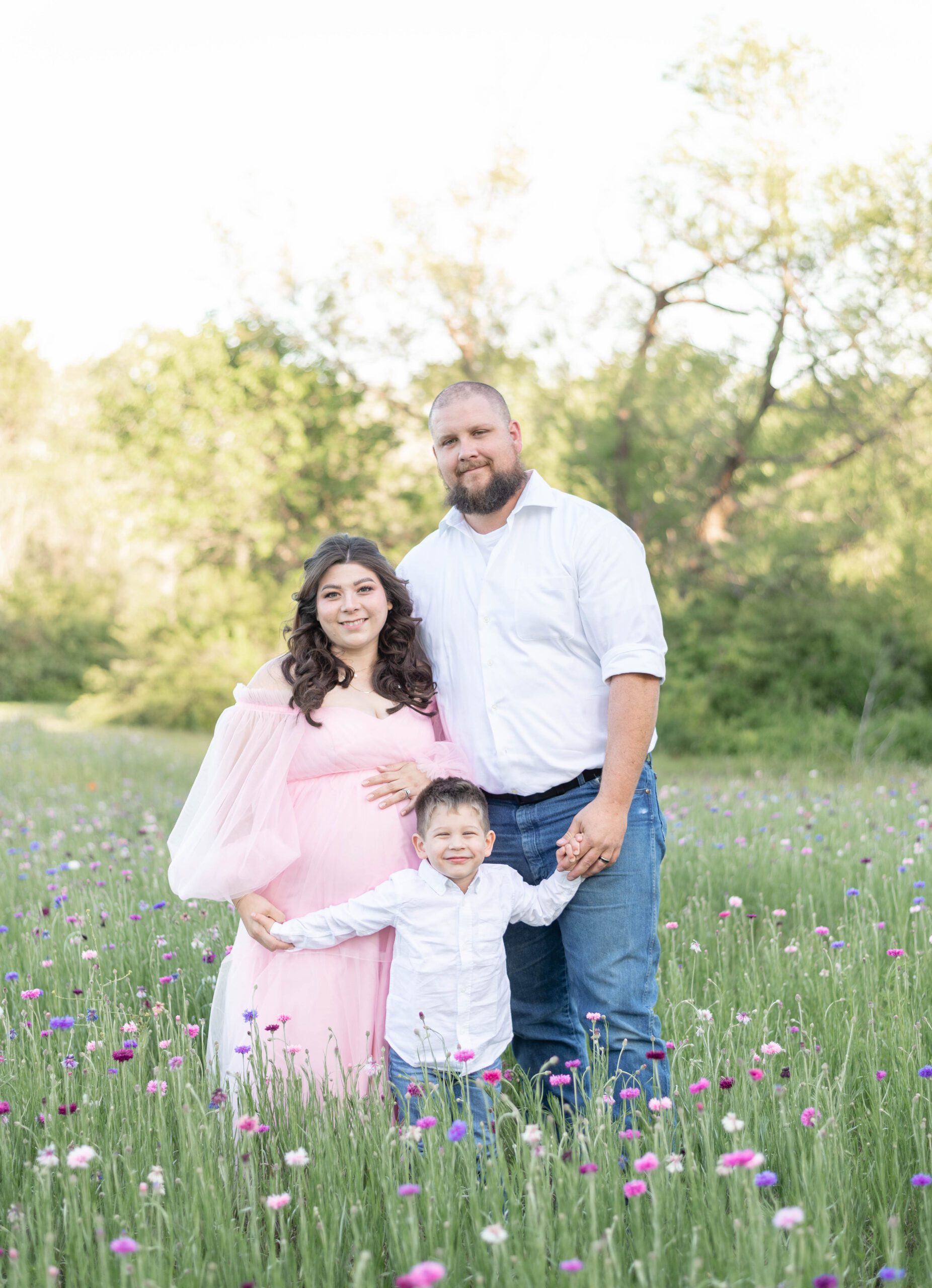 a family of three hold hands and smile toward the camera in a field of pink and purple wildflowers